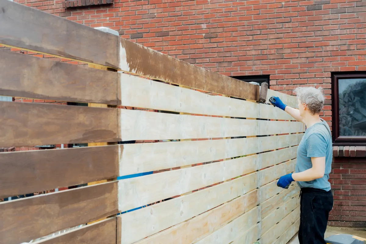 Crew member staining a horizontal-slat wood fence beside a brick home